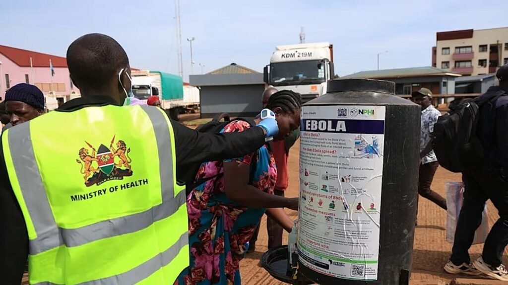 Kenyan Ministry of Health worker checks a traveler's temperature beside an Ebola information poster