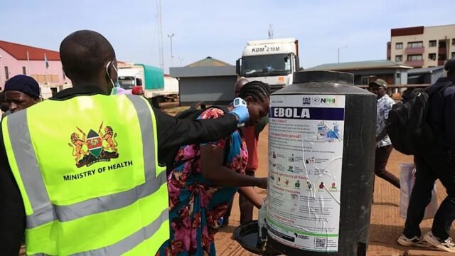 Kenyan Ministry of Health worker checks a traveler's temperature beside an Ebola information poster